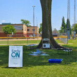 An EarlyON sign and outdoor programming is set up under a large tree beside a baseball diamond.
